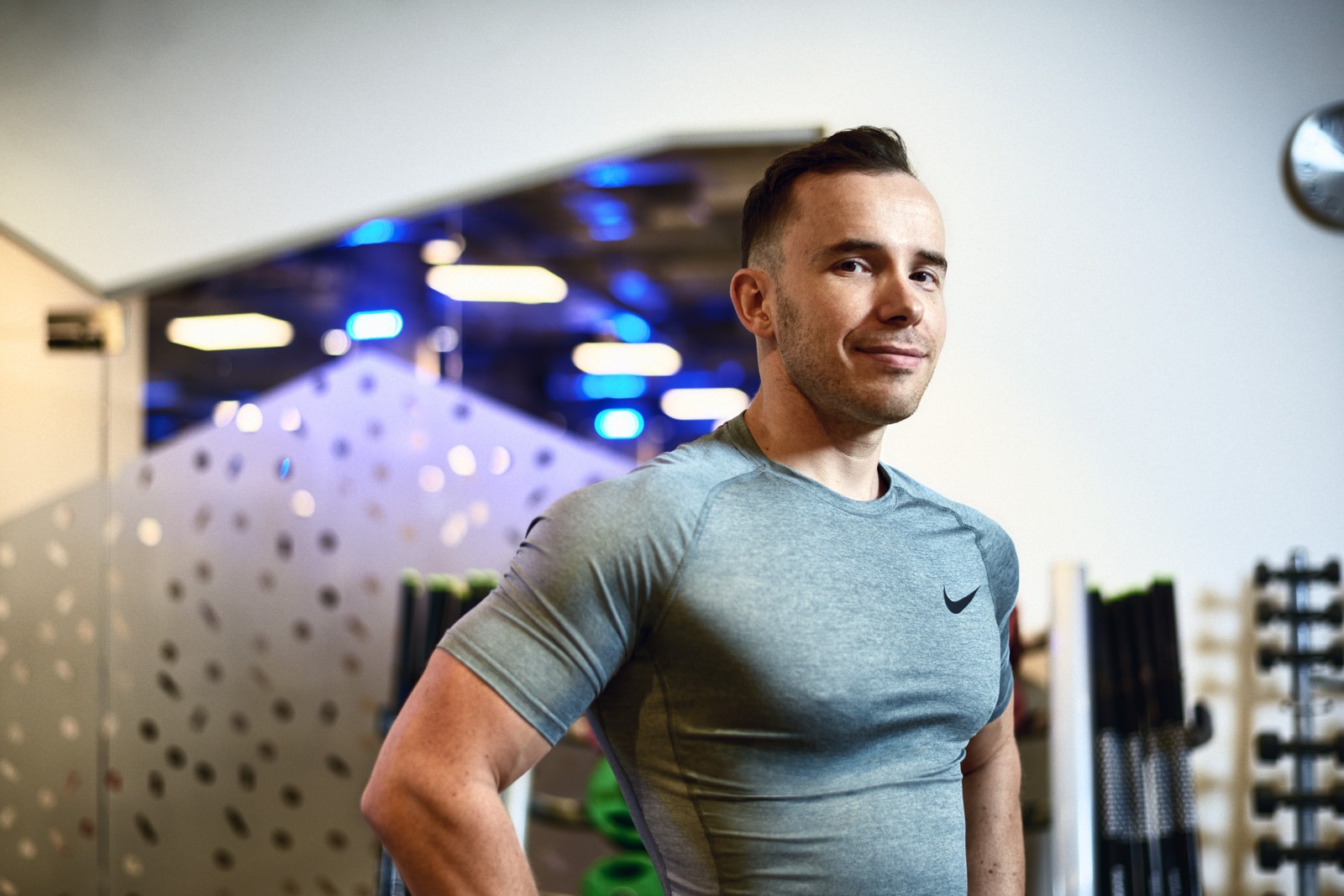 Uśmiechnięty mężczyzna w sportowej koszulce Nike pozuje w siłowni, w tle widoczne hantle i oświetlenie sufitowe.