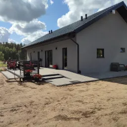 Parterowy dom z szarą elewacją i ciemną dachówką, widoczny taras z drewnianą balustradą i roślinami w donicach, trawnik w trakcie zakładania, błękitne niebo z chmurami w tle.