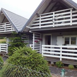 MagicHouse - Dwa identyczne domki letniskowe z białymi balustradami i szarą dachówką, widoczne zza metalowej siatki ogrodzeniowej.