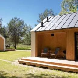 Dwa nowoczesne, drewniane domki na zielonej działce, jeden z zadaszonym tarasem z krzesłami i grillem, drugi z prostokątnymi oknami, błękitne niebo w tle.