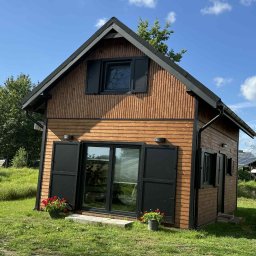 MagicHouse - Niewielki, parterowy dom z drewnianą elewacją w odcieniach brązu, z czarnymi okiennicami i dachem. Uroku dodają kwiaty w donicach przy wejściu.