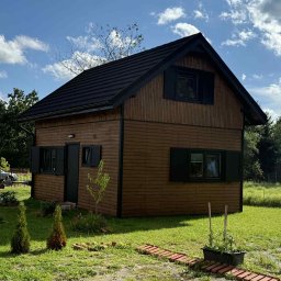 MagicHouse - Niewielki, dwukondygnacyjny dom z drewnianą elewacją i czarnym dachem, otoczony zielonym trawnikiem i młodymi drzewkami. Błękitne niebo z chmurami w tle.