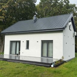 MagicHouse - Nowoczesny, parterowy dom z ciemną dachówką i tarasem. Biała elewacja, duże okna przesuwne. Otoczenie: trawnik i drzewa. Widoczny klimatyzator na ścianie.