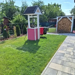 Ogród w Dębnicy Kaszubskiej z dekoracyjną studnią w kolorze różowo-białym, trawnikiem, ścieżką z kostki brukowej i drewutnią w tle, wykonany w słoneczny dzień.