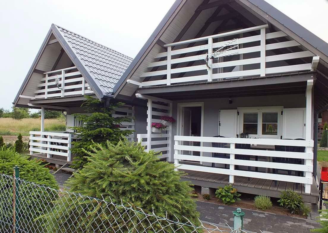 Dwa identyczne domki letniskowe z białymi balustradami i szarą dachówką, widoczne zza metalowej siatki ogrodzeniowej.