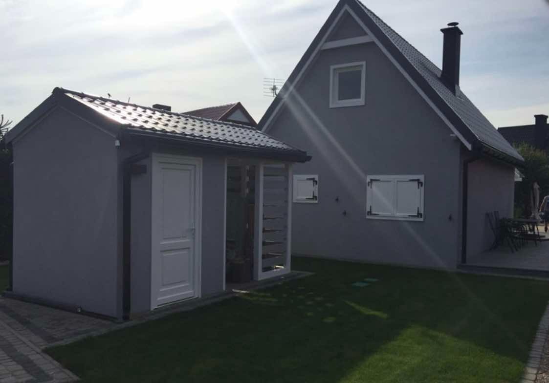 Nowoczesny dom z ciemnoszarą elewacją i białymi okiennicami, obok znajduje się mały budynek gospodarczy z przeszklonym wejściem, całość otoczona trawnikiem.