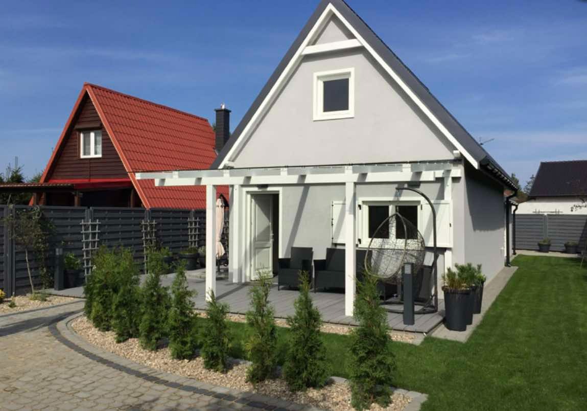 Nowoczesny dom z szarą elewacją, białą stolarką okienną i pergolą nad tarasem, otoczony zielonym trawnikiem i ozdobnymi krzewami.