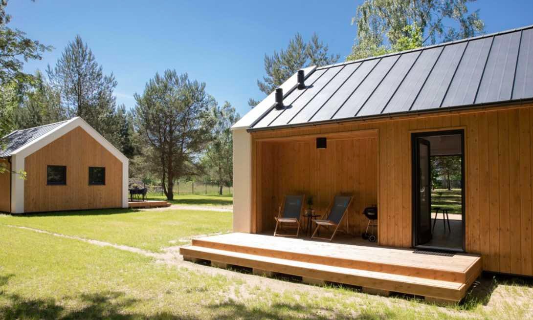 Dwa nowoczesne, drewniane domki na zielonej działce, jeden z zadaszonym tarasem z krzesłami i grillem, drugi z prostokątnymi oknami, błękitne niebo w tle.