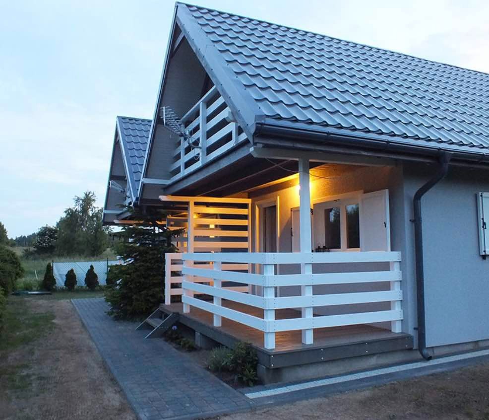 Dwa identyczne domki z szarą elewacją i ciemnoszarą dachówką, białe balustrady werand, widoczne anteny na dachach, oświetlenie werand, kostka brukowa prowadząca do wejścia.