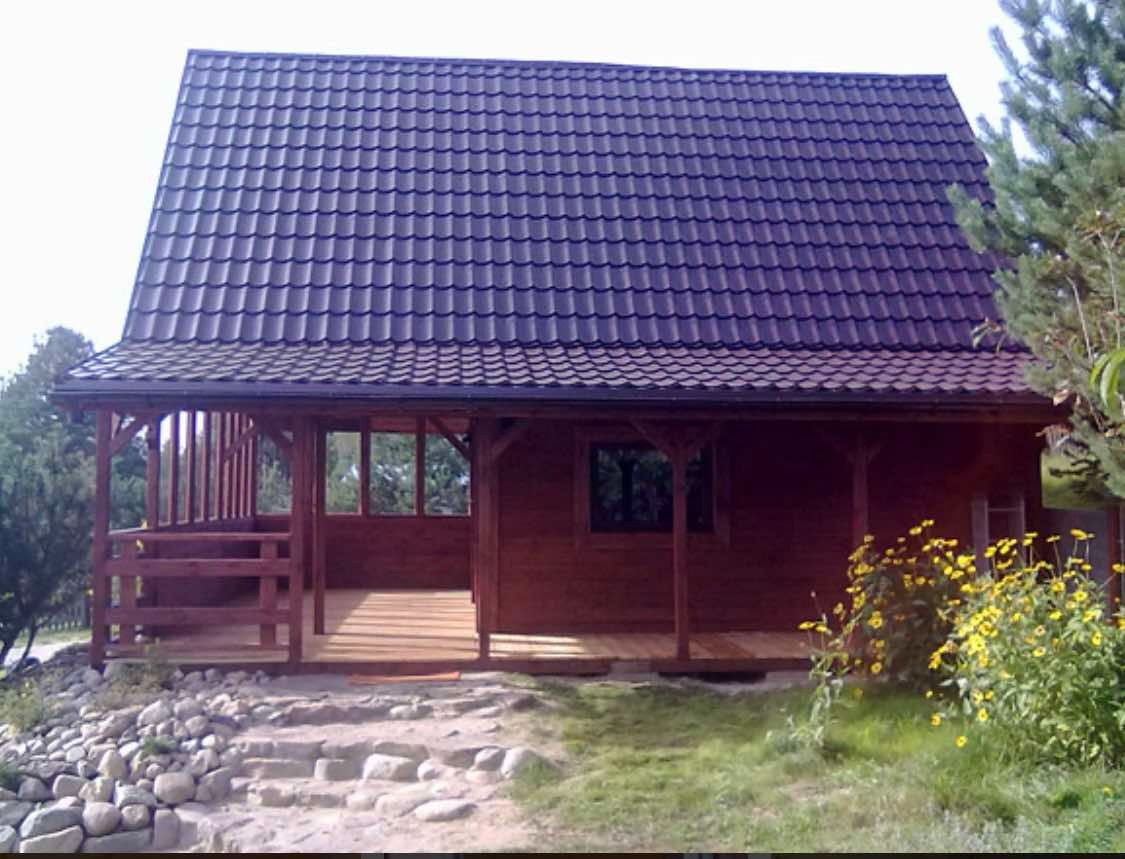 Drewniany dom z zadaszonym tarasem i ciemną dachówką, zlokalizowany w Dębnicy Kaszubskiej, otoczony zielenią i kamieniami.