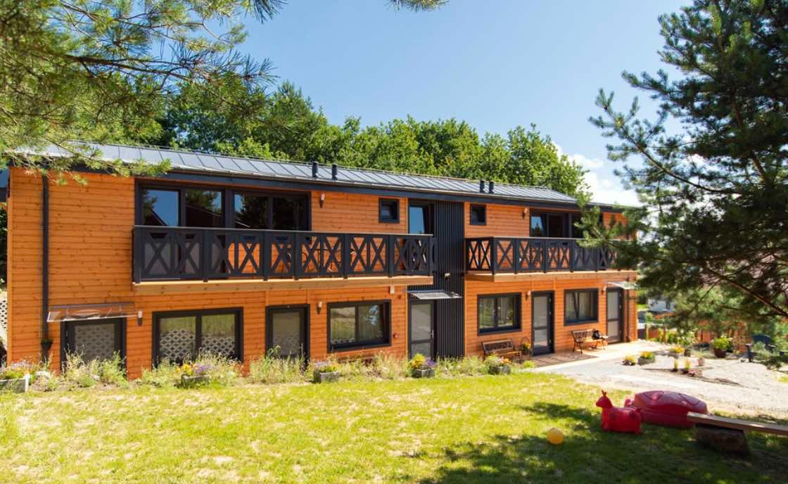 Dwukondygnacyjny budynek z drewnianą elewacją w kolorze pomarańczowym, z czarnymi balustradami balkonów i ramami okiennymi, otoczony zielonym trawnikiem i drzewami.