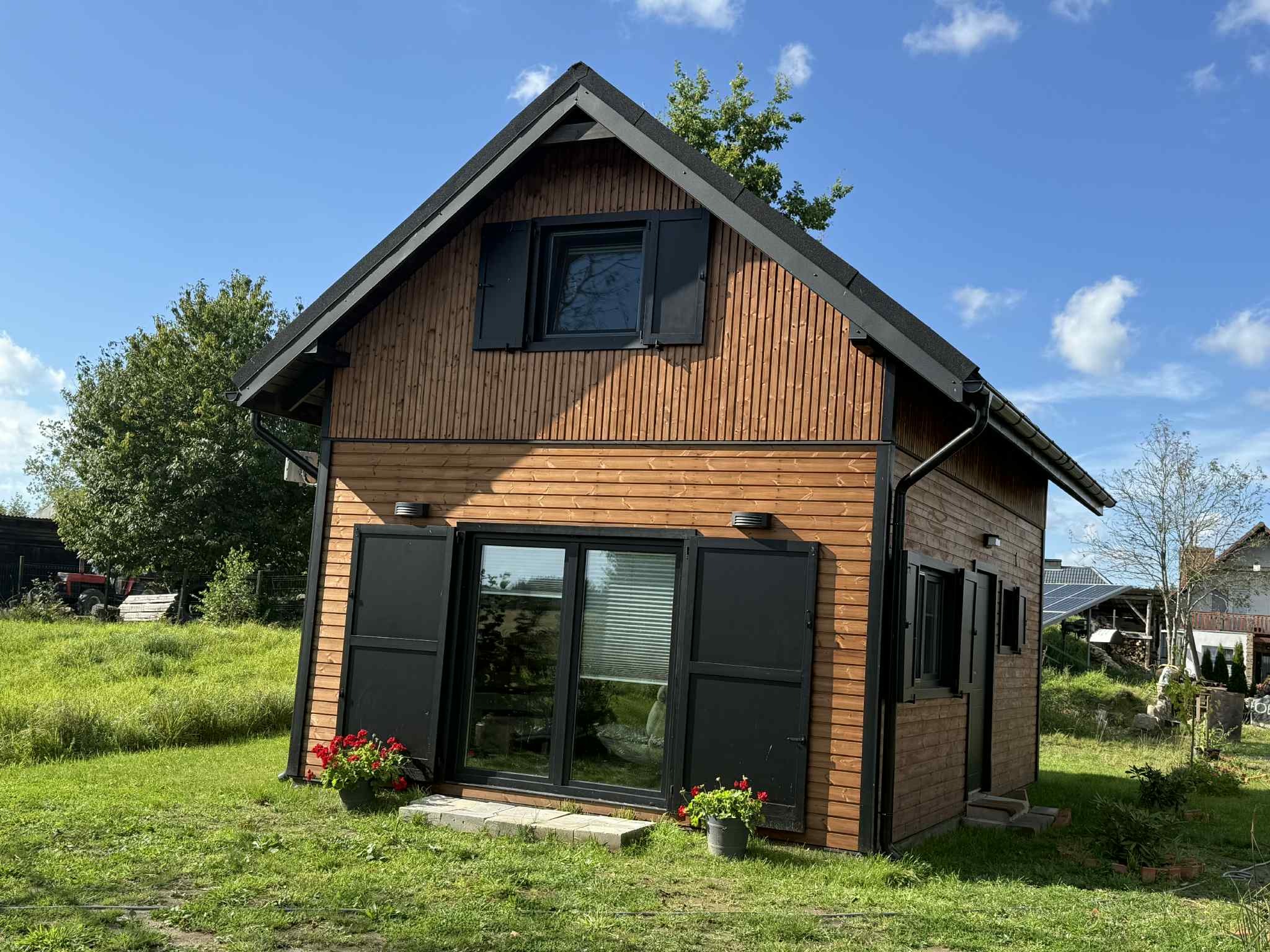 Niewielki, parterowy dom z drewnianą elewacją w odcieniach brązu, z czarnymi okiennicami i dachem. Uroku dodają kwiaty w donicach przy wejściu.