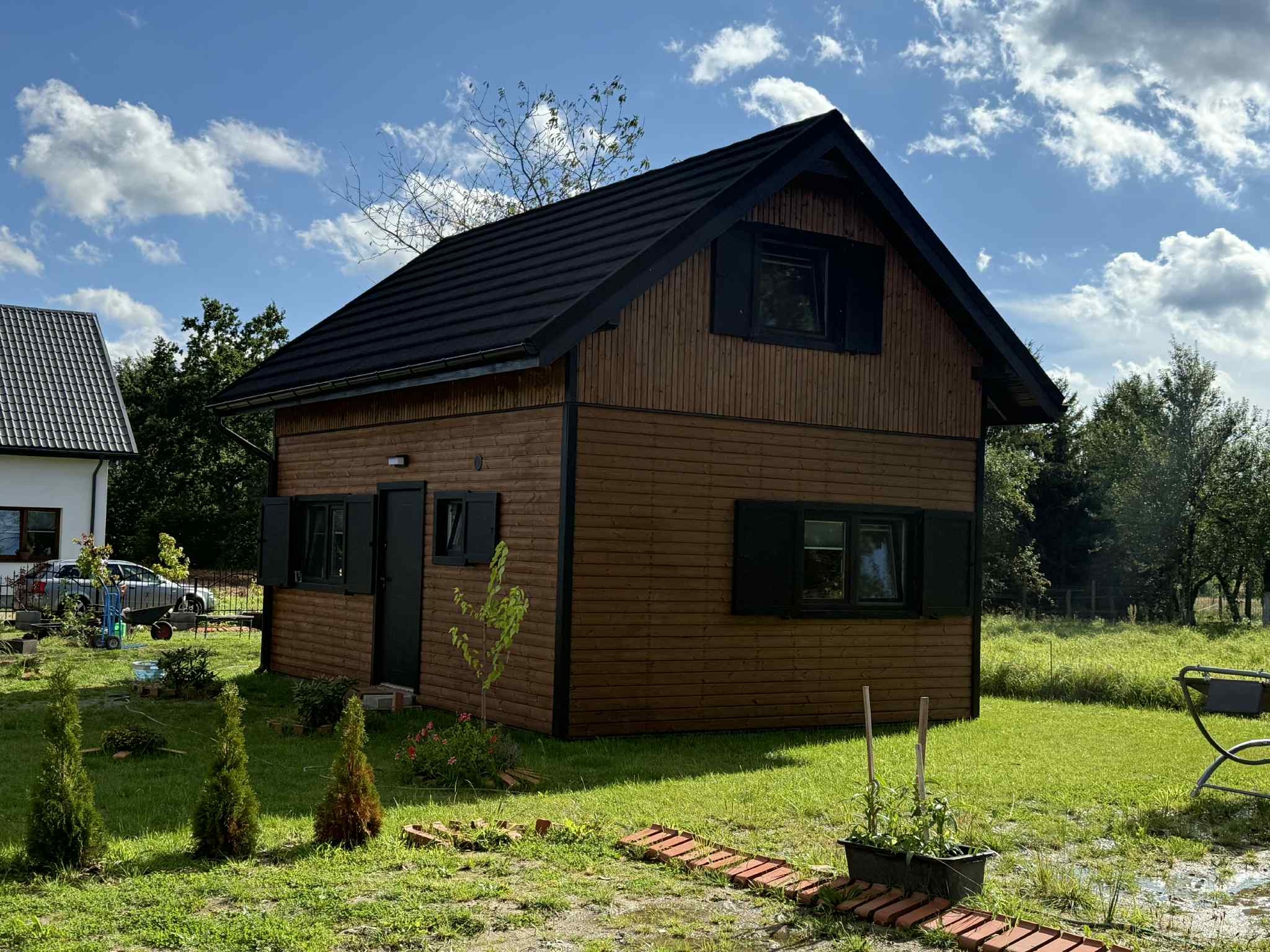 Niewielki, dwukondygnacyjny dom z drewnianą elewacją i czarnym dachem, otoczony zielonym trawnikiem i młodymi drzewkami. Błękitne niebo z chmurami w tle.
