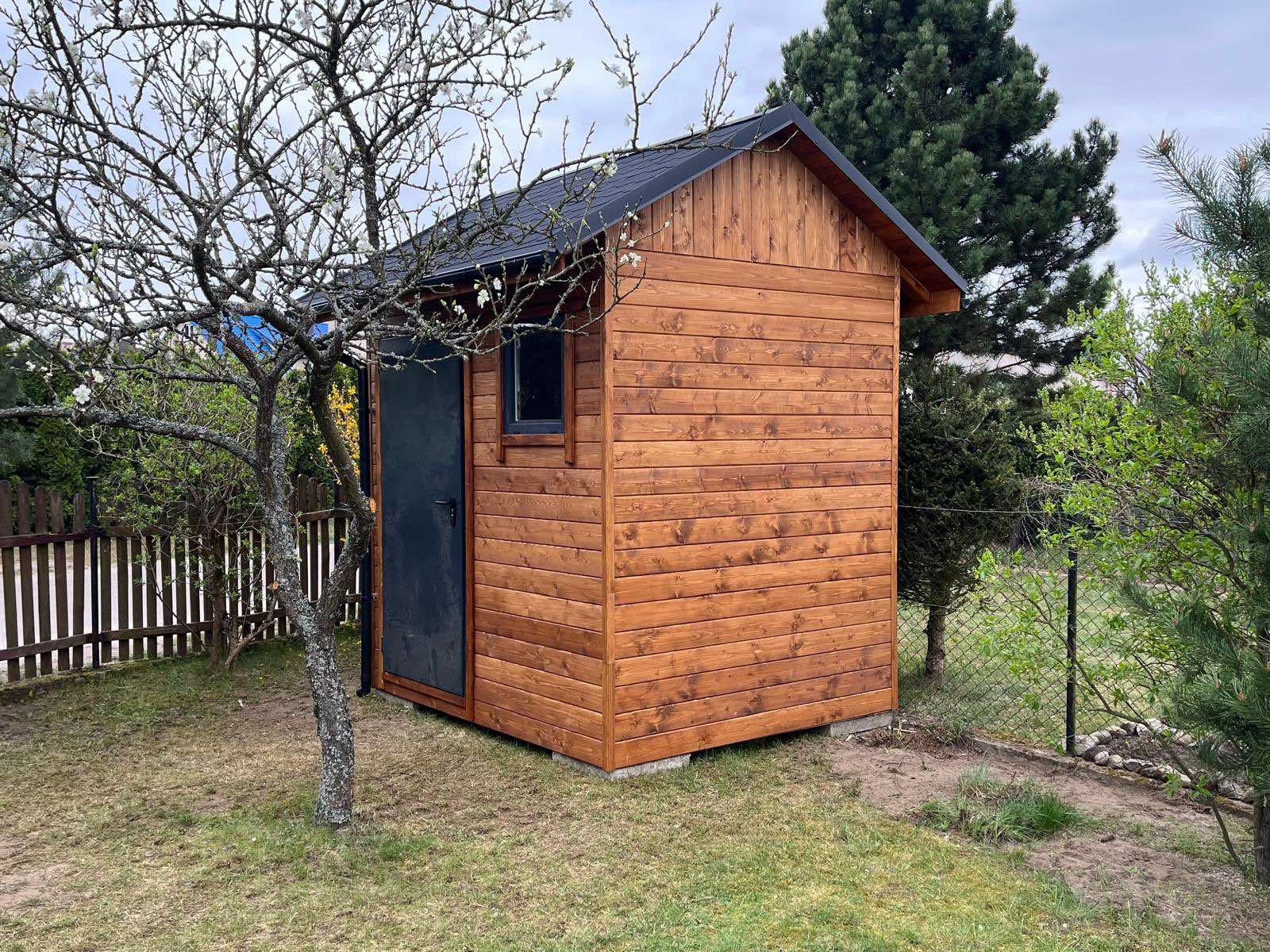 Drewniana altana ogrodowa z czarnym dachem i drzwiami, widoczna z boku na tle trawnika i drzew. Konstrukcja na betonowych bloczkach, z widoczną fakturą drewna.