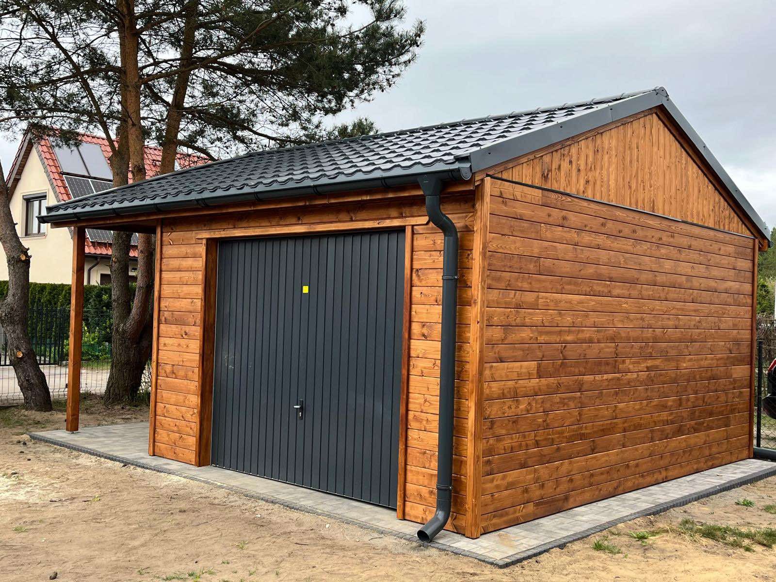 Drewniany garaż z szarą bramą segmentową i dachem z blachodachówki, zadaszenie z boku, widoczne rynny, kostka brukowa wokół, drzewa w tle.