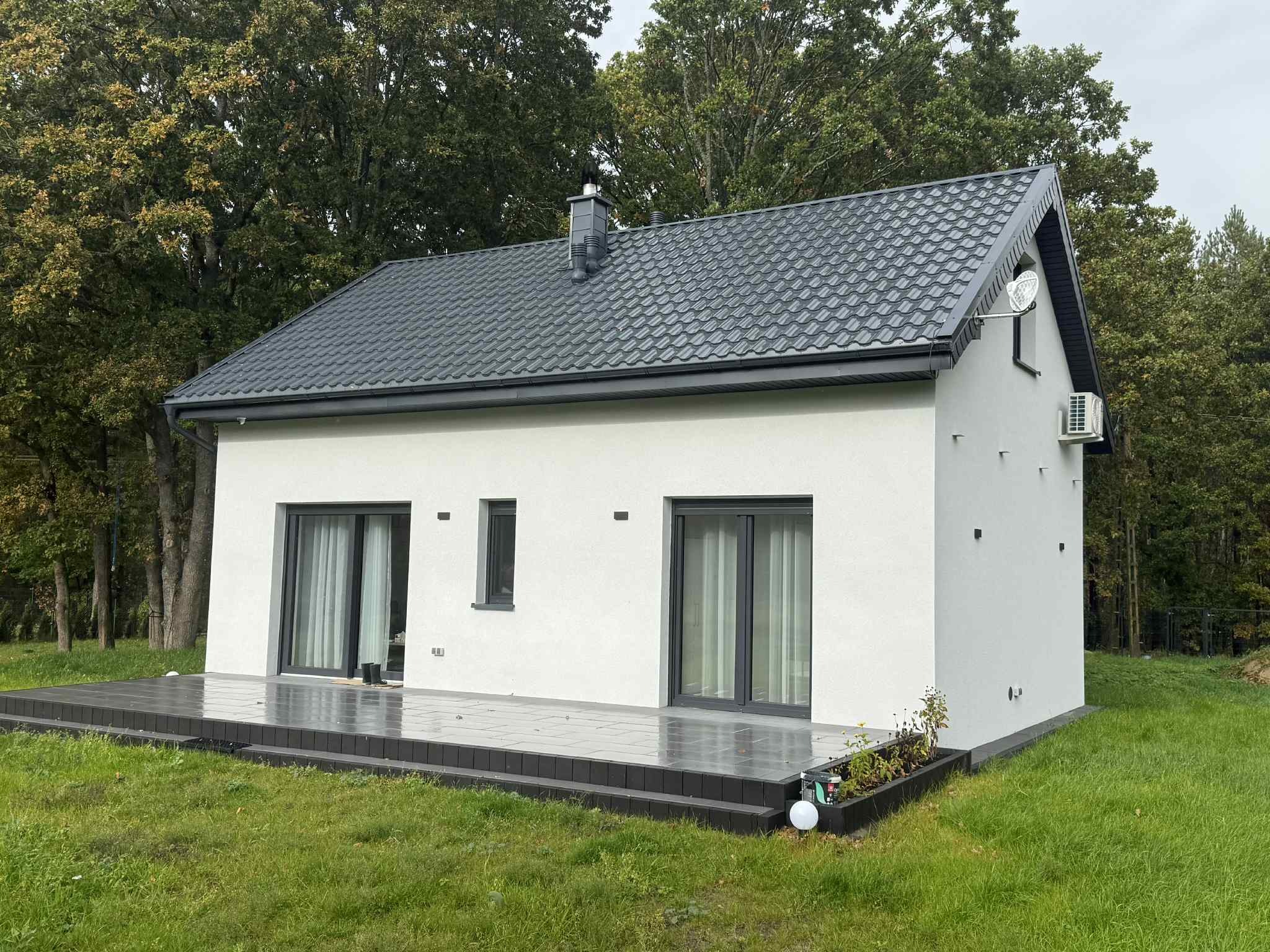 Nowoczesny, parterowy dom z ciemną dachówką i tarasem. Biała elewacja, duże okna przesuwne. Otoczenie: trawnik i drzewa. Widoczny klimatyzator na ścianie.