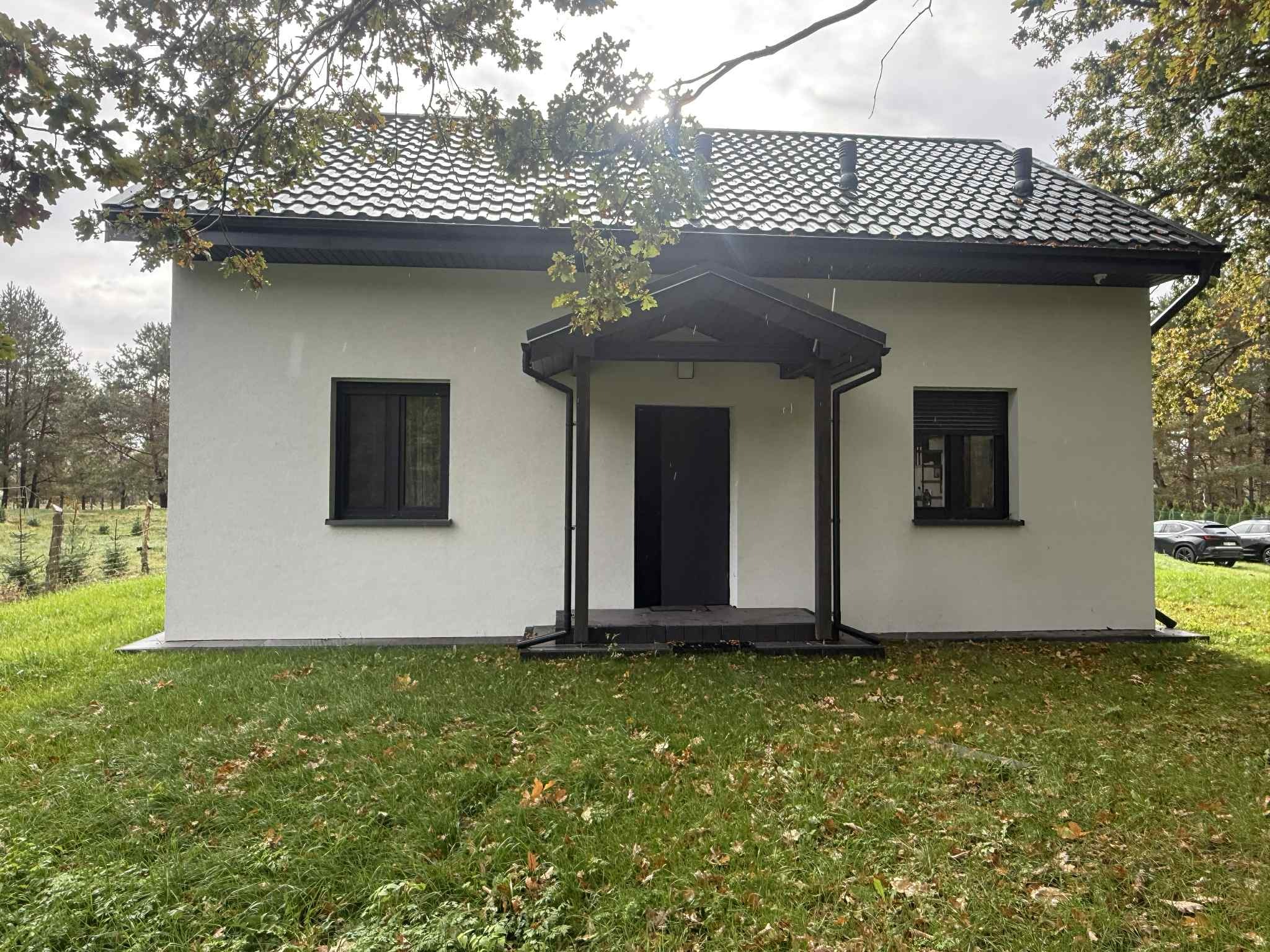 Biały dom z czarnym dachem i oknami, otoczony zielenią. Widoczny fragment drzewa w rogu kadru. Minimalistyczny styl budynku.