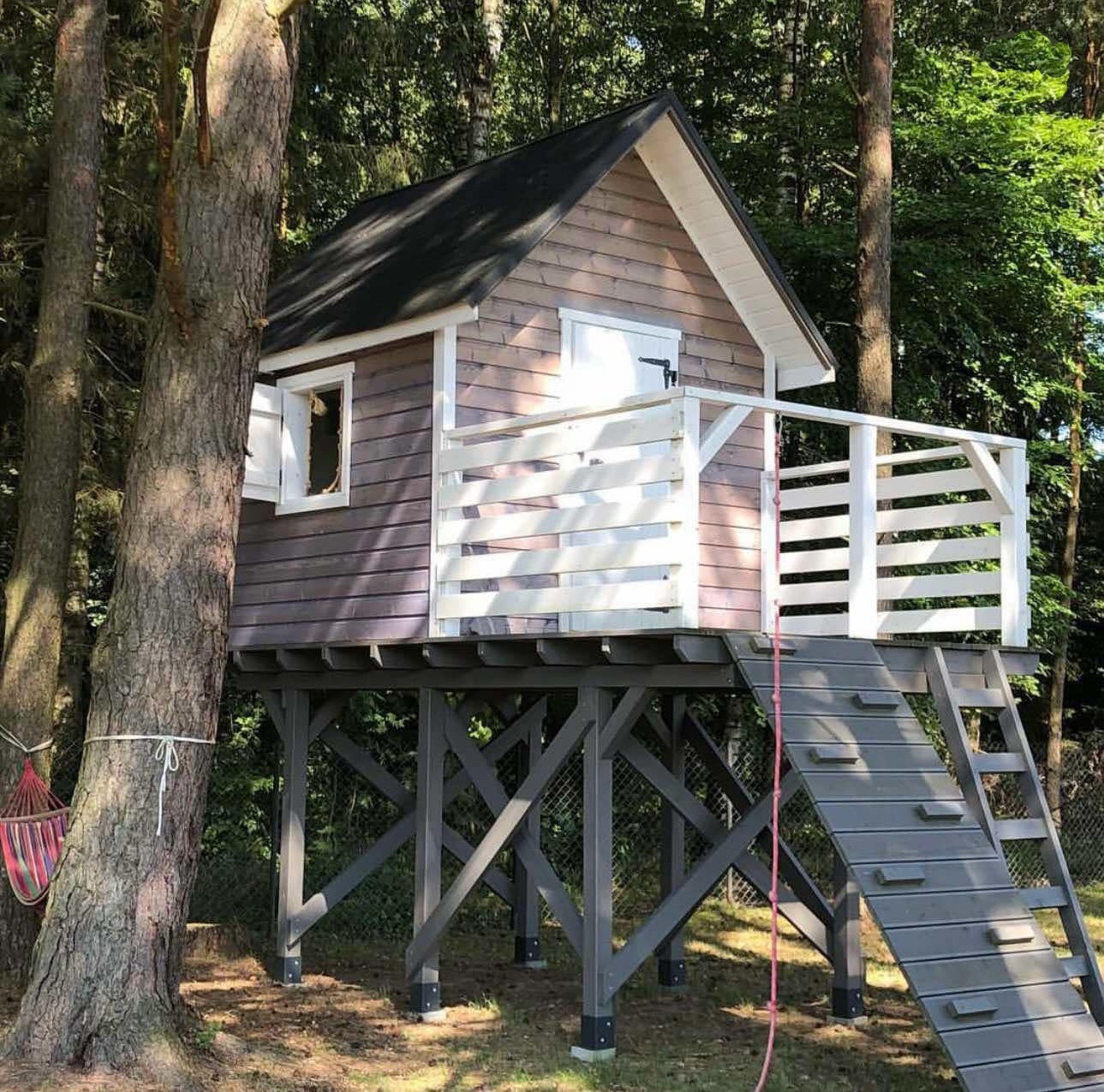 Drewniany domek dla dzieci na drzewie, z małym balkonem i schodami, w otoczeniu drzew. Konstrukcja na podwyższeniu, pomalowana w odcieniach szarości i bieli.