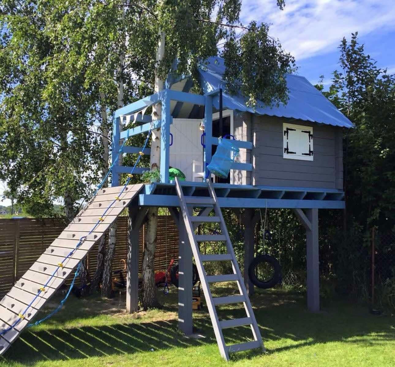 Drewniany plac zabaw z domkiem na drzewie w Dębnicy Kaszubskiej. Konstrukcja pomalowana na szaro i niebiesko, z drabinką sznurową i oponą.