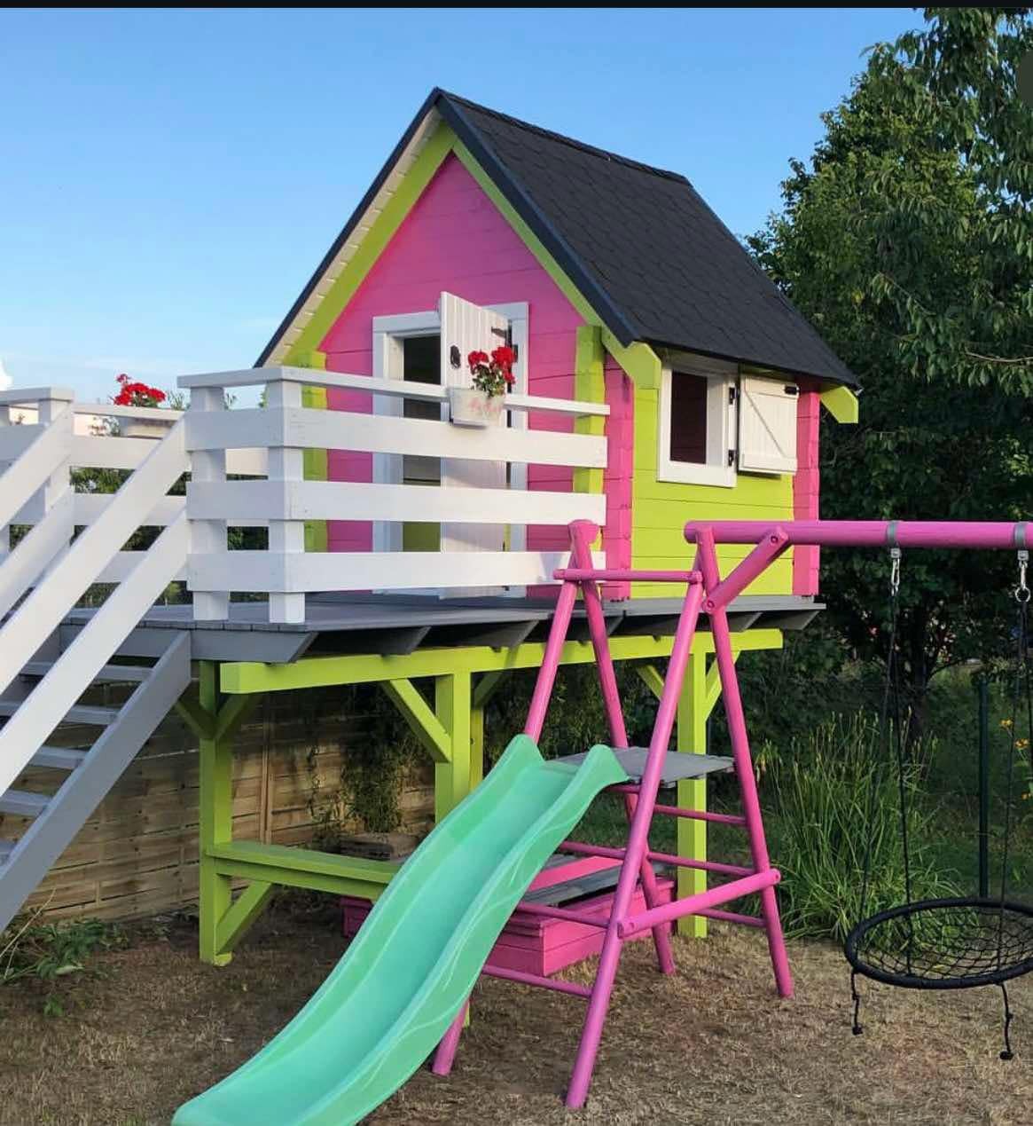 Kolorowa altana dla dzieci z huśtawką i zjeżdżalnią, pomalowana na różowo i zielono, z białymi elementami. Na balkonie altany czerwone kwiaty w doniczkach.
