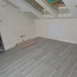 Krystian salach - Pokój na poddaszu w trakcie prac wykończeniowych: jasnoszare panele podłogowe, przygotowane ściany do malowania, okno dachowe z folią ochronną, widoczne puszki elektryczne.