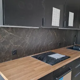 Nowoczesna kuchnia z czarnymi szafkami, blatem imitującym drewno i marmurowym panelem ściennym w trakcie montażu, widoczne ślady po naklejkach na szafkach.