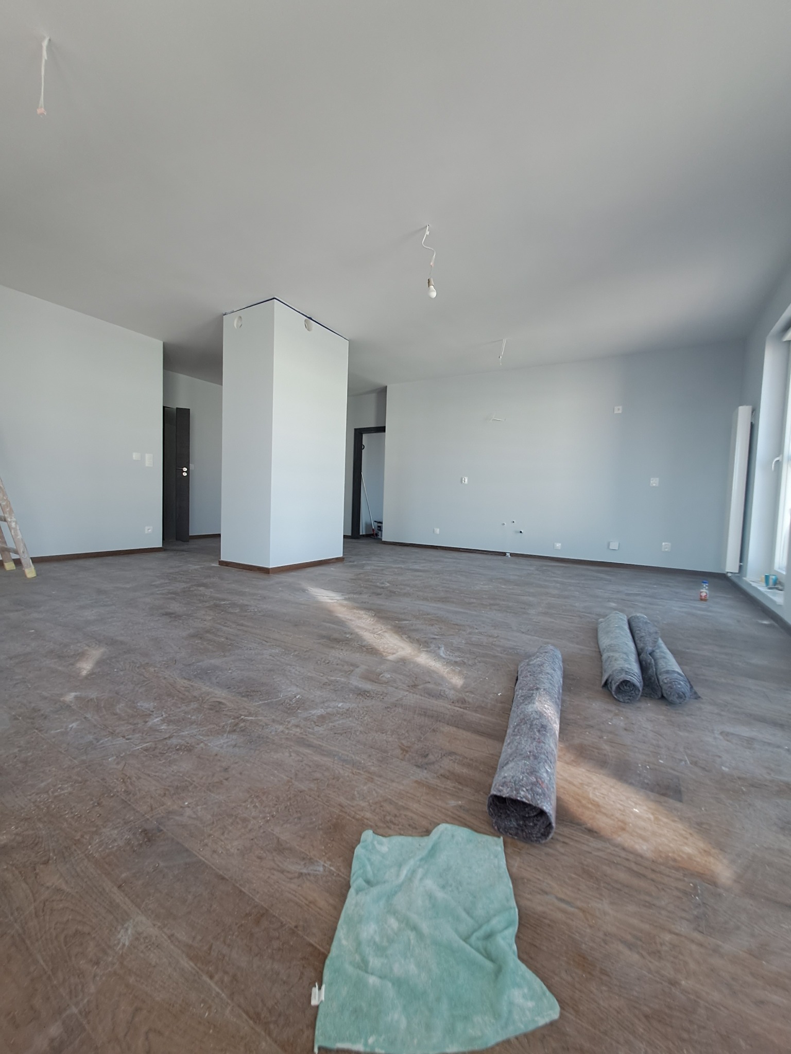 Przygotowanie do prac wykończeniowych w przestronnym, jasnym wnętrzu: zabezpieczona podłoga drewniana, zwinięte maty ochronne, ślady po remoncie na podłodze i świeżo pomalowane ściany w odcieniach...