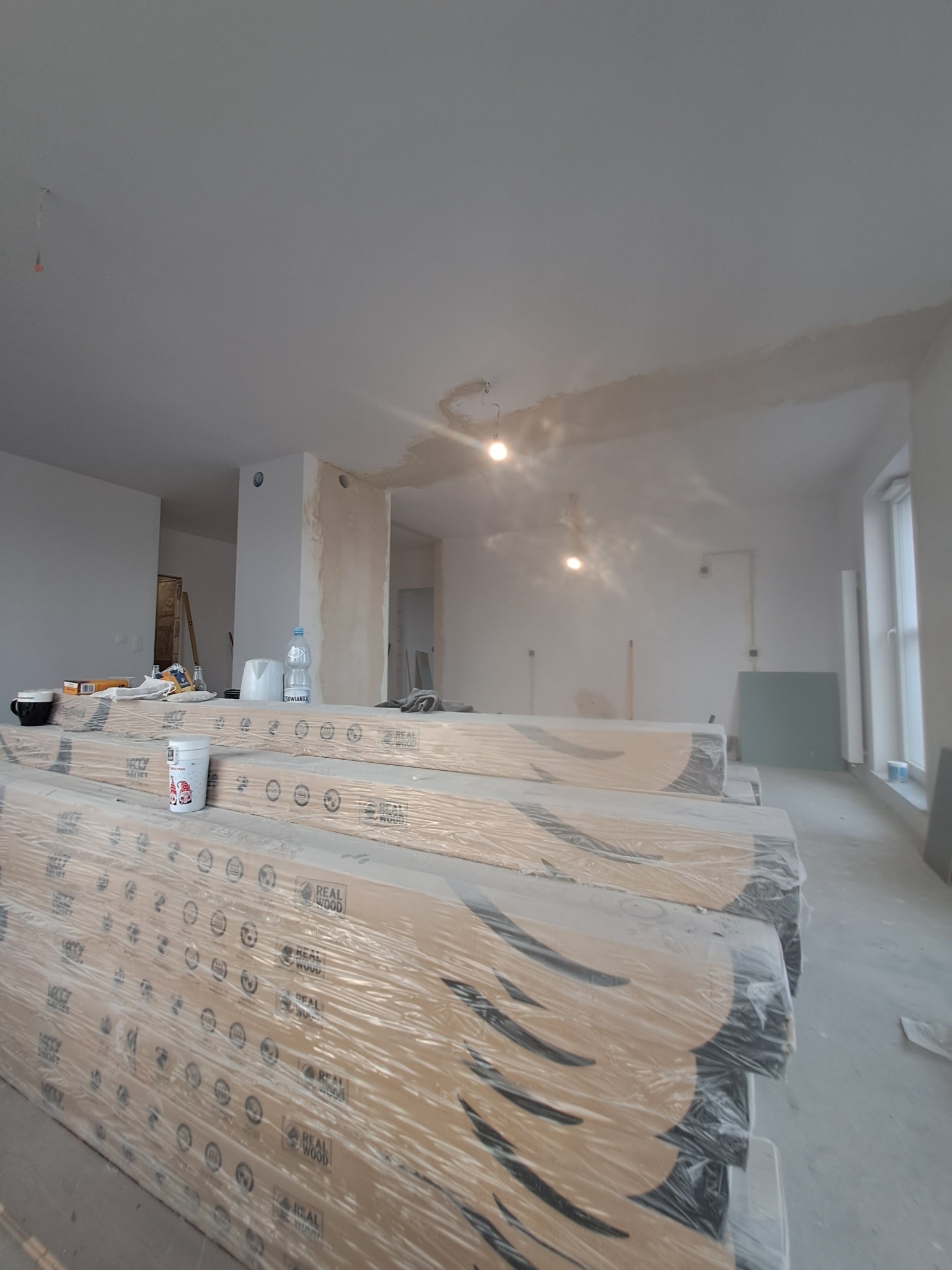 Stosy zapakowanych paneli podłogowych z widocznym logo 'Real Wood' w surowym wnętrzu remontowanego pomieszczenia z niedokończonymi gładziami na ścianach i suficie.