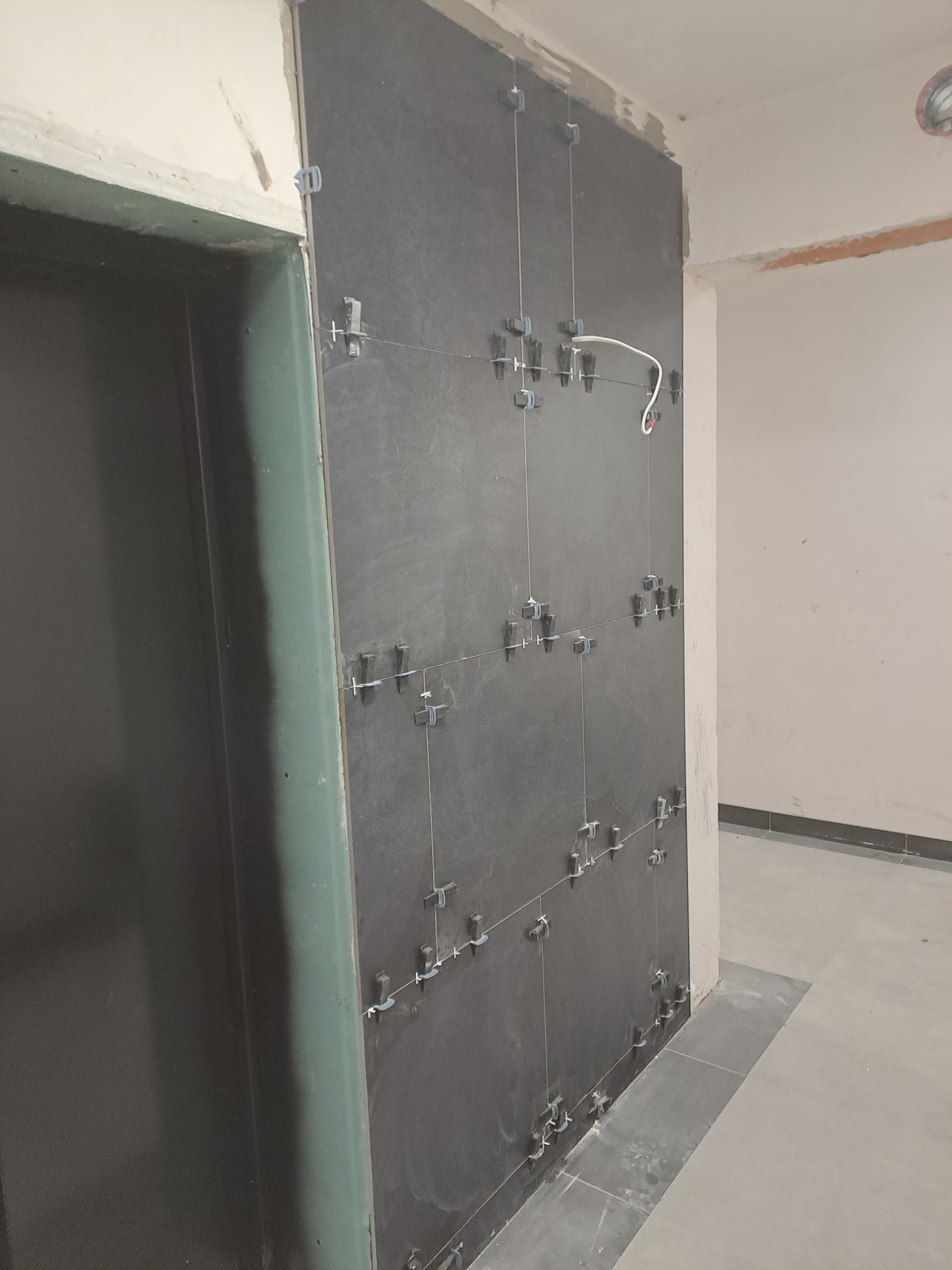 Montaż ciemnych, kwadratowych płytek ściennych z użyciem krzyżyków dystansowych i sznurków poziomujących w trakcie remontu pomieszczenia.