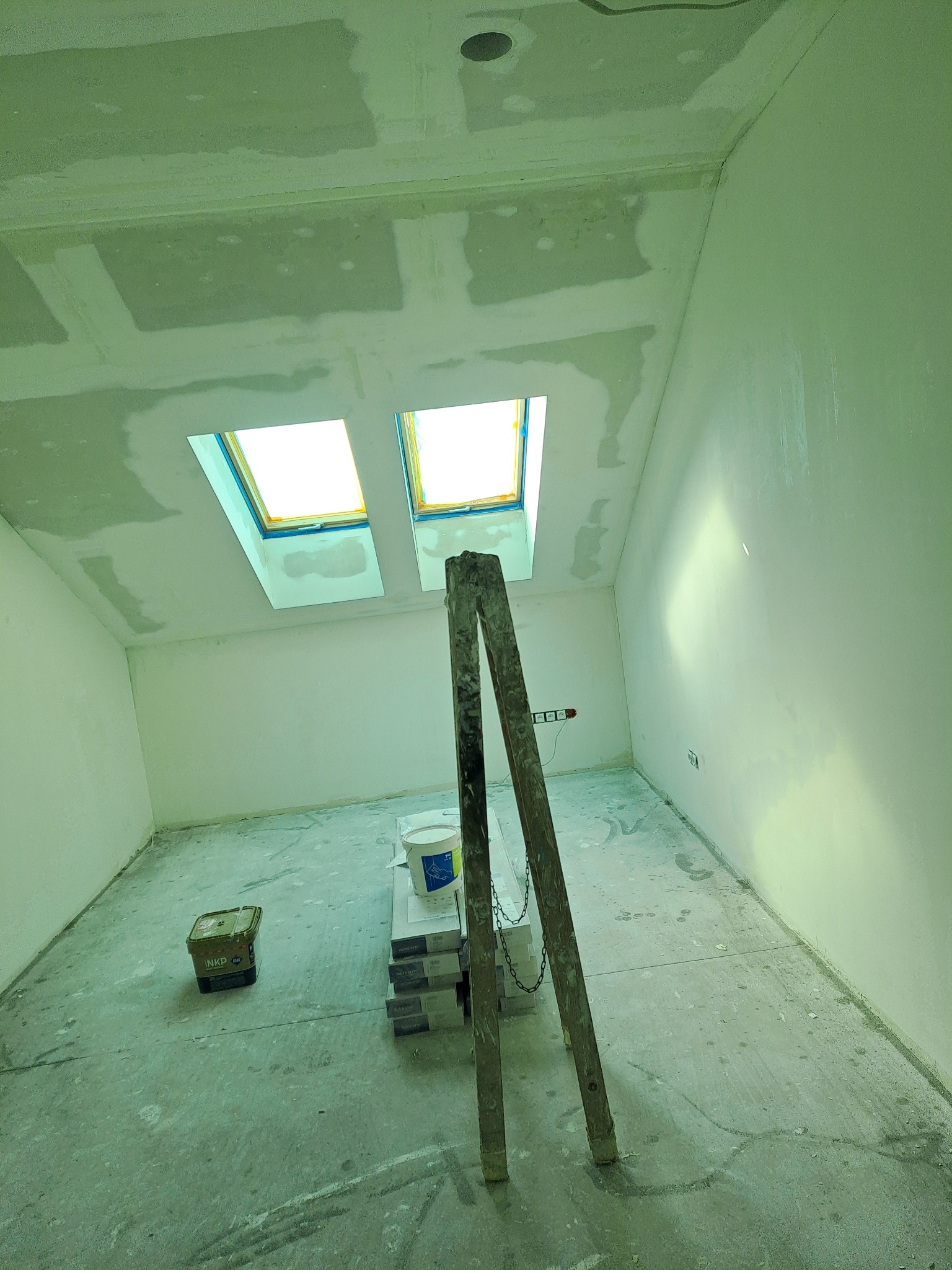 Świeżo szpachlowane poddasze z dwoma oknami dachowymi, widoczna drabina, puszka z farbą i paczki paneli podłogowych, przygotowane do dalszych prac wykończeniowych.