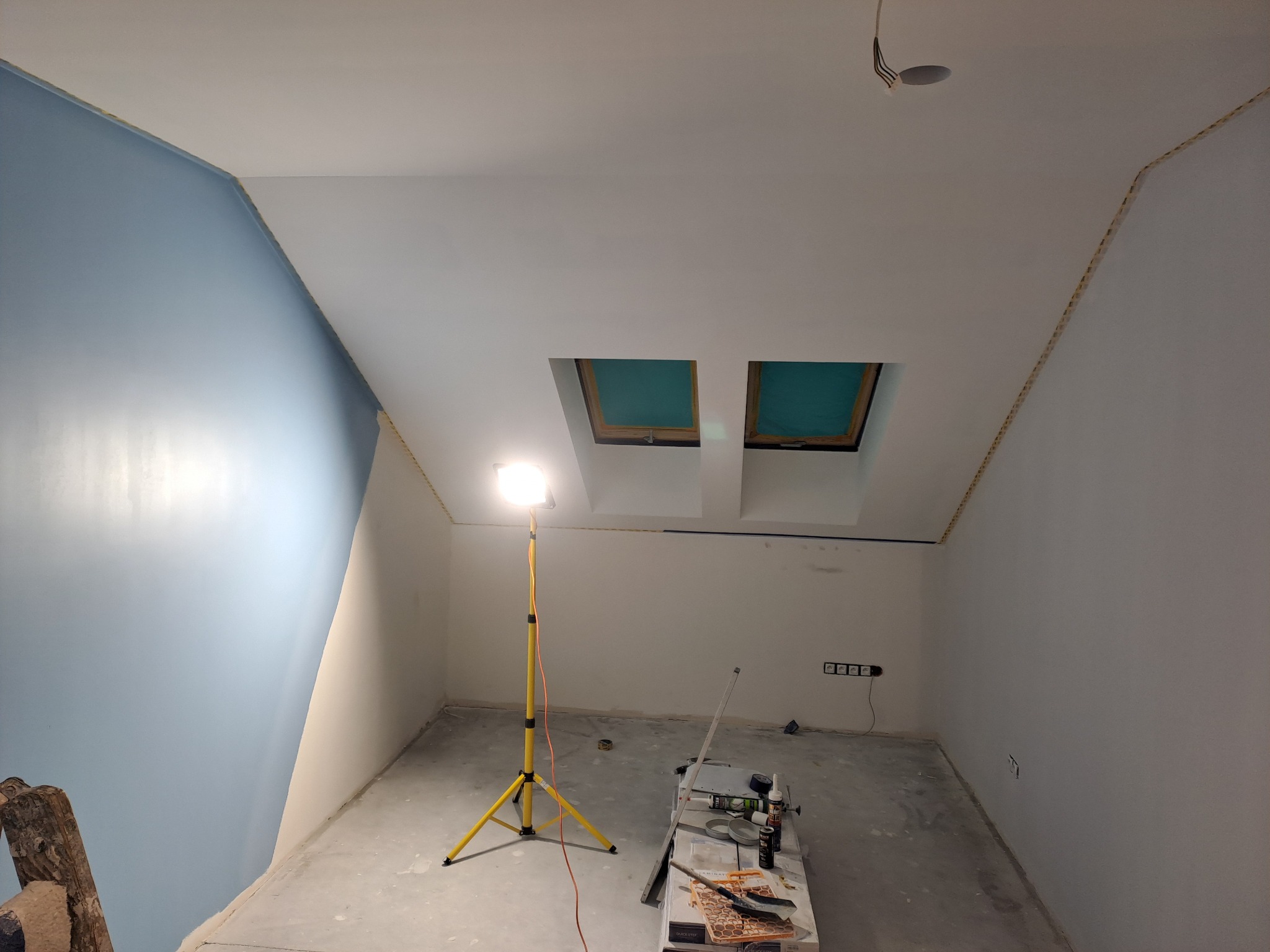 Pokój na poddaszu w trakcie remontu: pomalowana na błękitno ściana, biały sufit z dwoma oknami dachowymi, reflektor na statywie i narzędzia malarskie na podłodze.