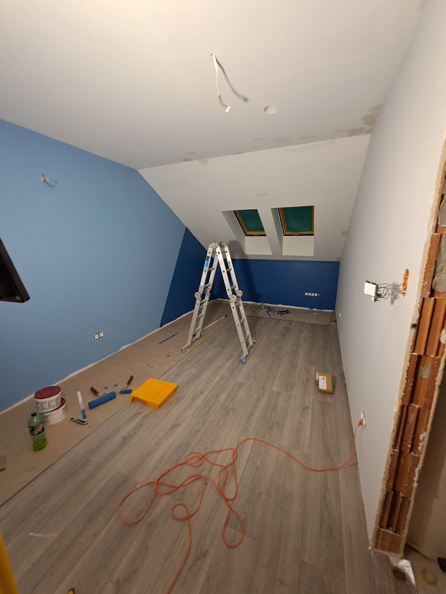 Pokój na poddaszu w trakcie malowania: ściana pomalowana na niebiesko do połowy wysokości, reszta ścian na biało, rozstawiona drabina aluminiowa, akcesoria malarskie na podłodze zabezpieczonej...