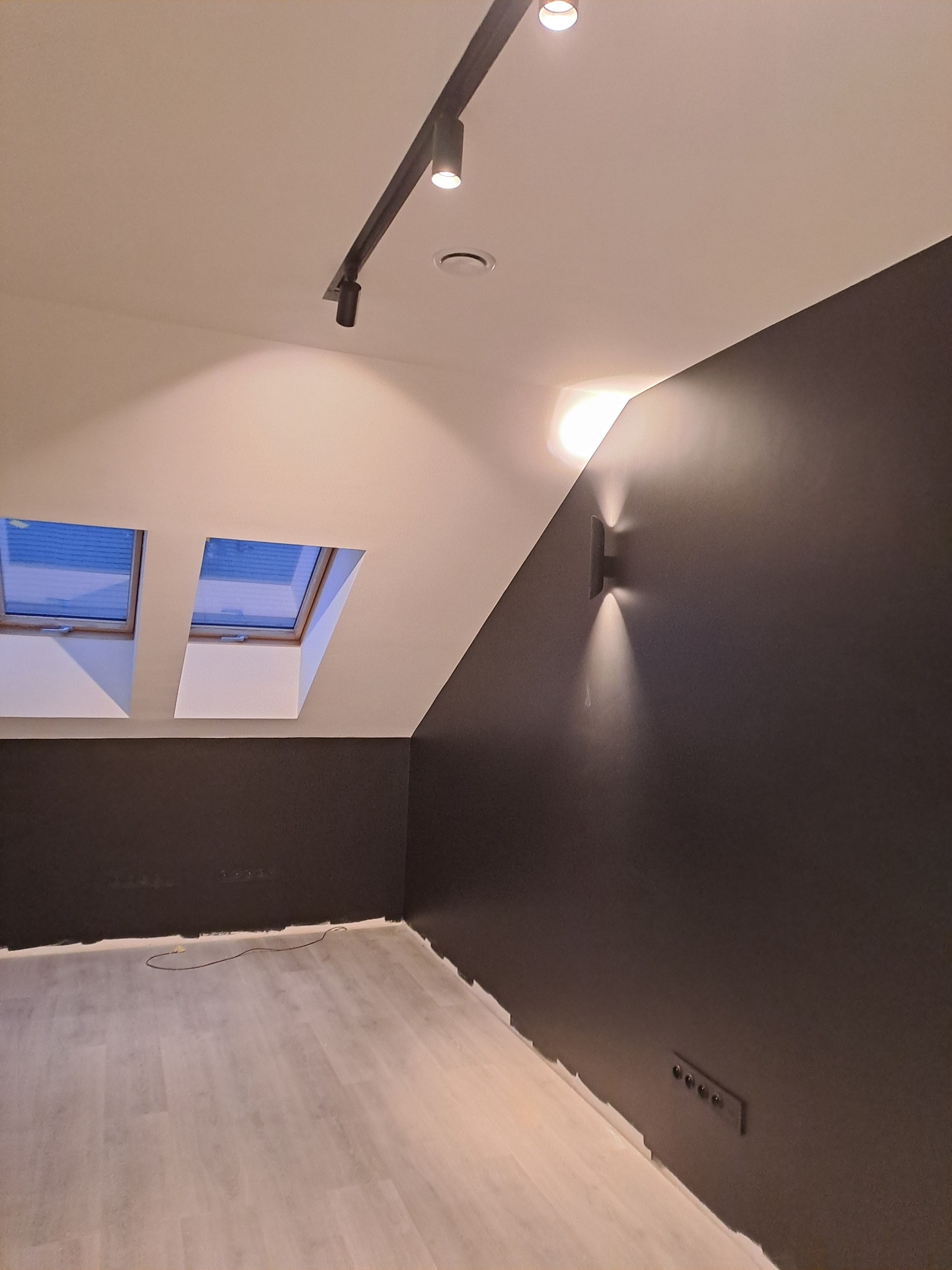 Pomieszczenie na poddaszu po remoncie: szara ściana, biały sufit ze skośnym oknem dachowym, oświetlenie szynowe i kinkiet.