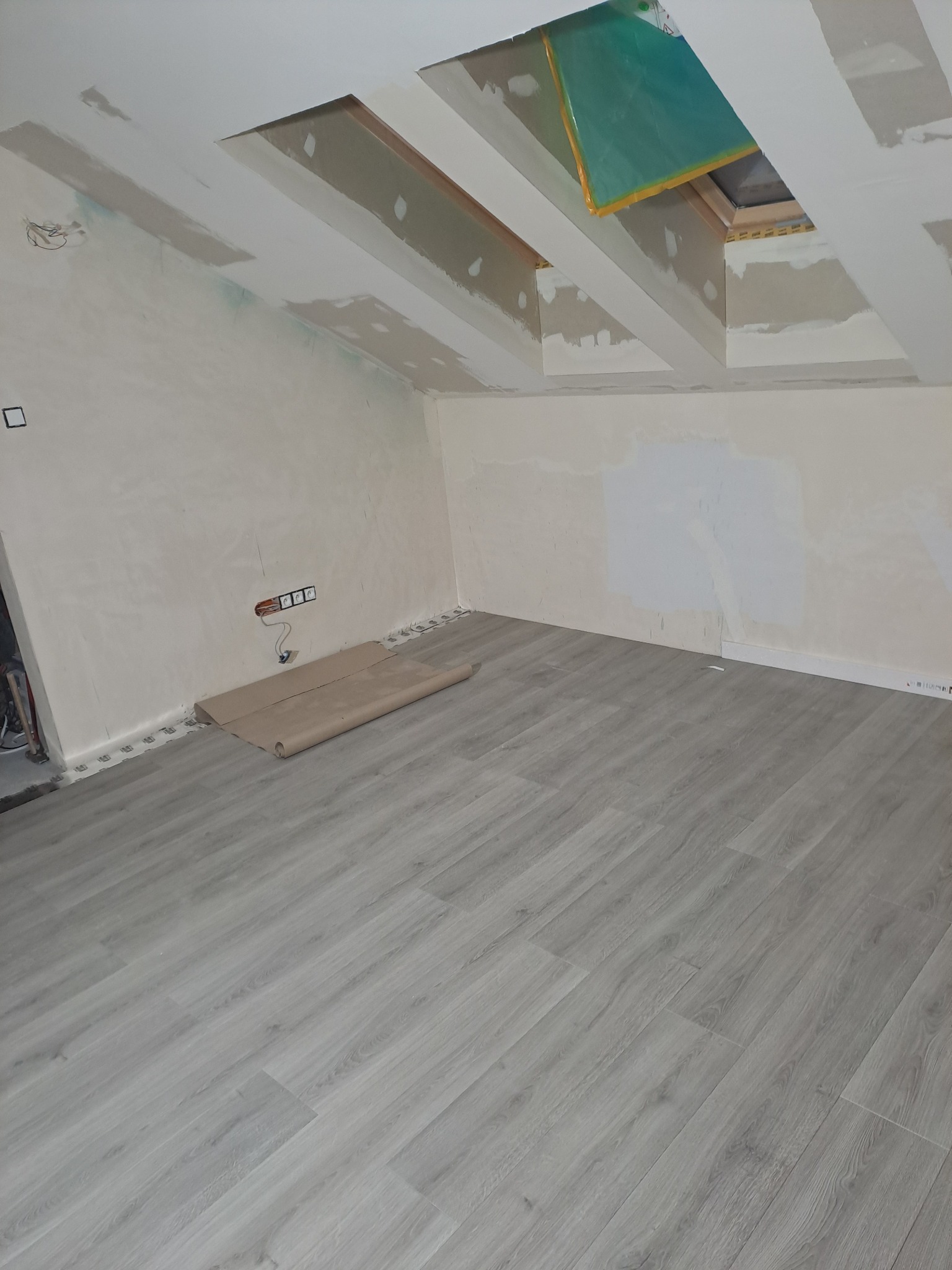 Pokój na poddaszu w trakcie prac wykończeniowych: jasnoszare panele podłogowe, przygotowane ściany do malowania, okno dachowe z folią ochronną, widoczne puszki elektryczne.