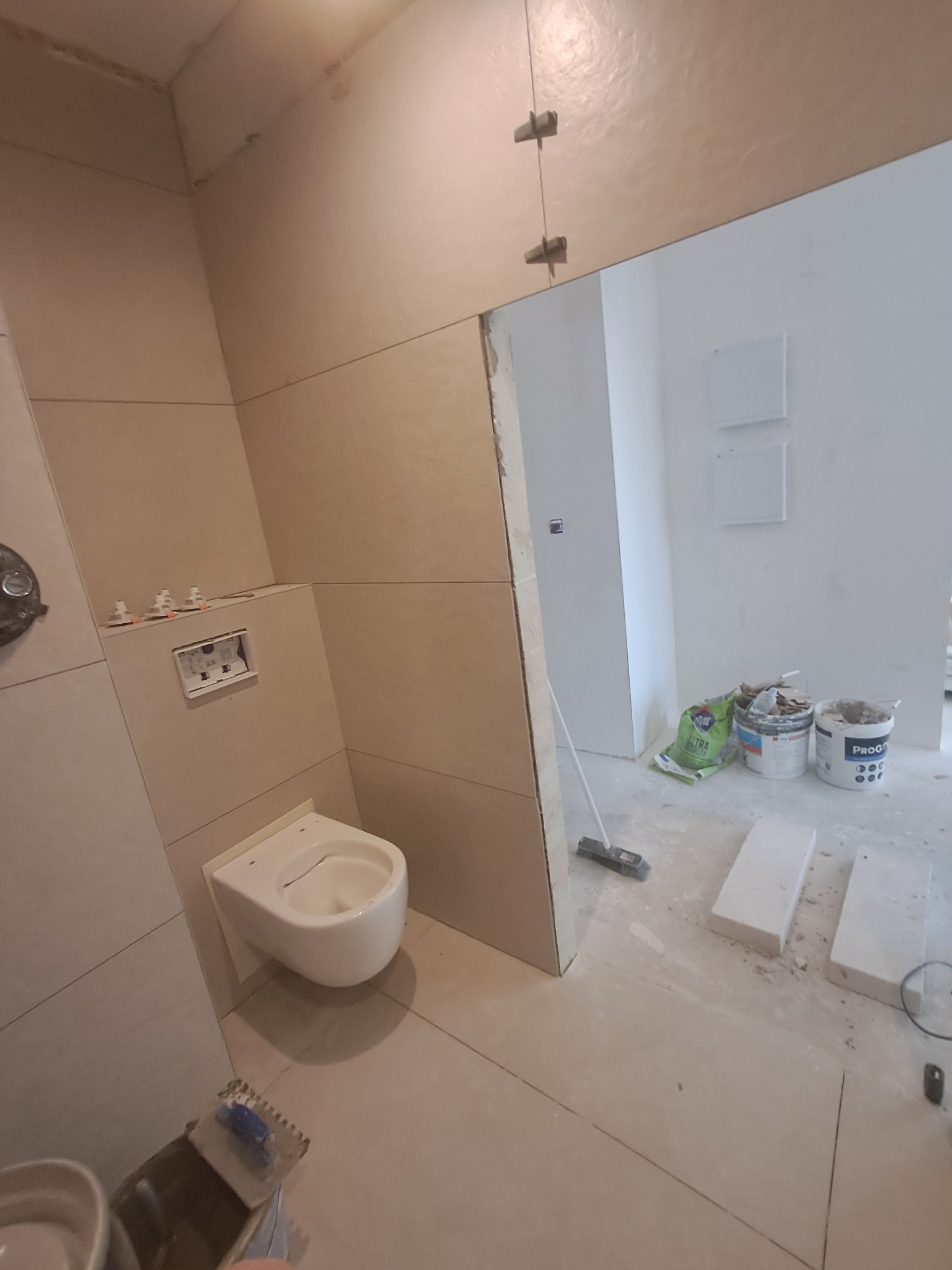 Łazienka w trakcie wykańczania: zamontowana podwieszana toaleta, widoczne płytki na ścianach, worki i pojemniki z materiałami budowlanymi w przejściu do innego pomieszczenia, krzyżyki dystansowe...