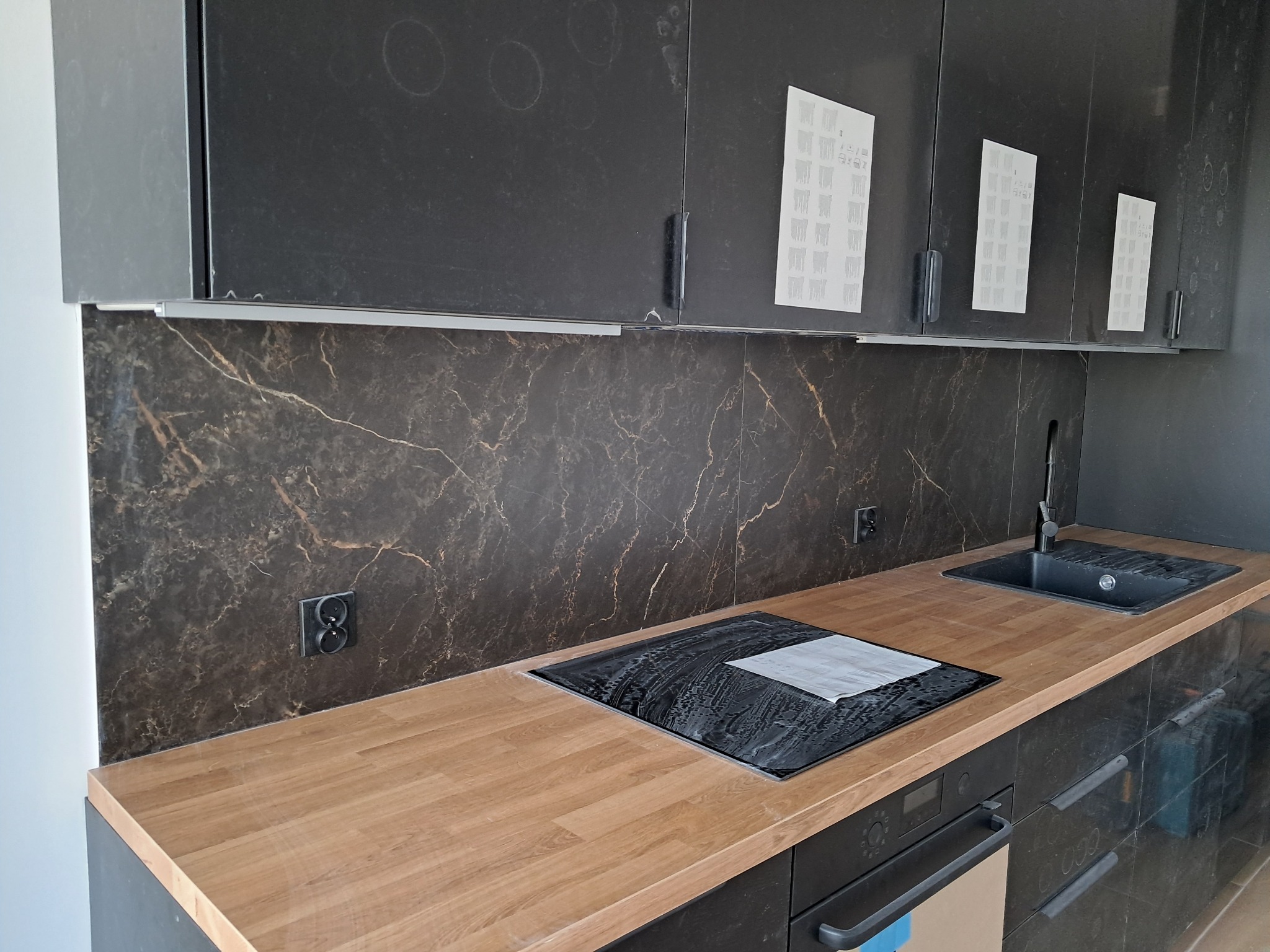 Nowoczesna kuchnia z czarnymi szafkami, blatem imitującym drewno i marmurowym panelem ściennym w trakcie montażu, widoczne ślady po naklejkach na szafkach.