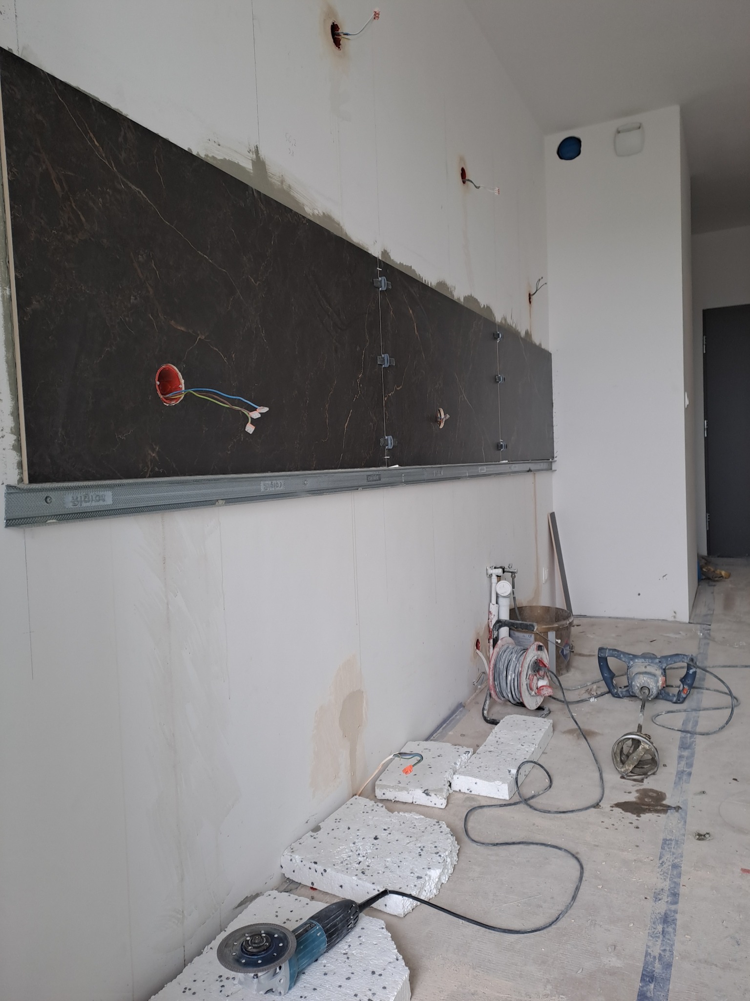 Montaż płytek imitujących marmur na ścianie, widoczne narzędzia i akcesoria glazurnicze, w tle wystające kable elektryczne