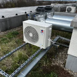 Biała jednostka zewnętrzna klimatyzacji marki Gree zamontowana na metalowej konstrukcji wsporczej na dachu budynku, widoczne rury wentylacyjne i inne urządzenia klimatyzacyjne w tle.