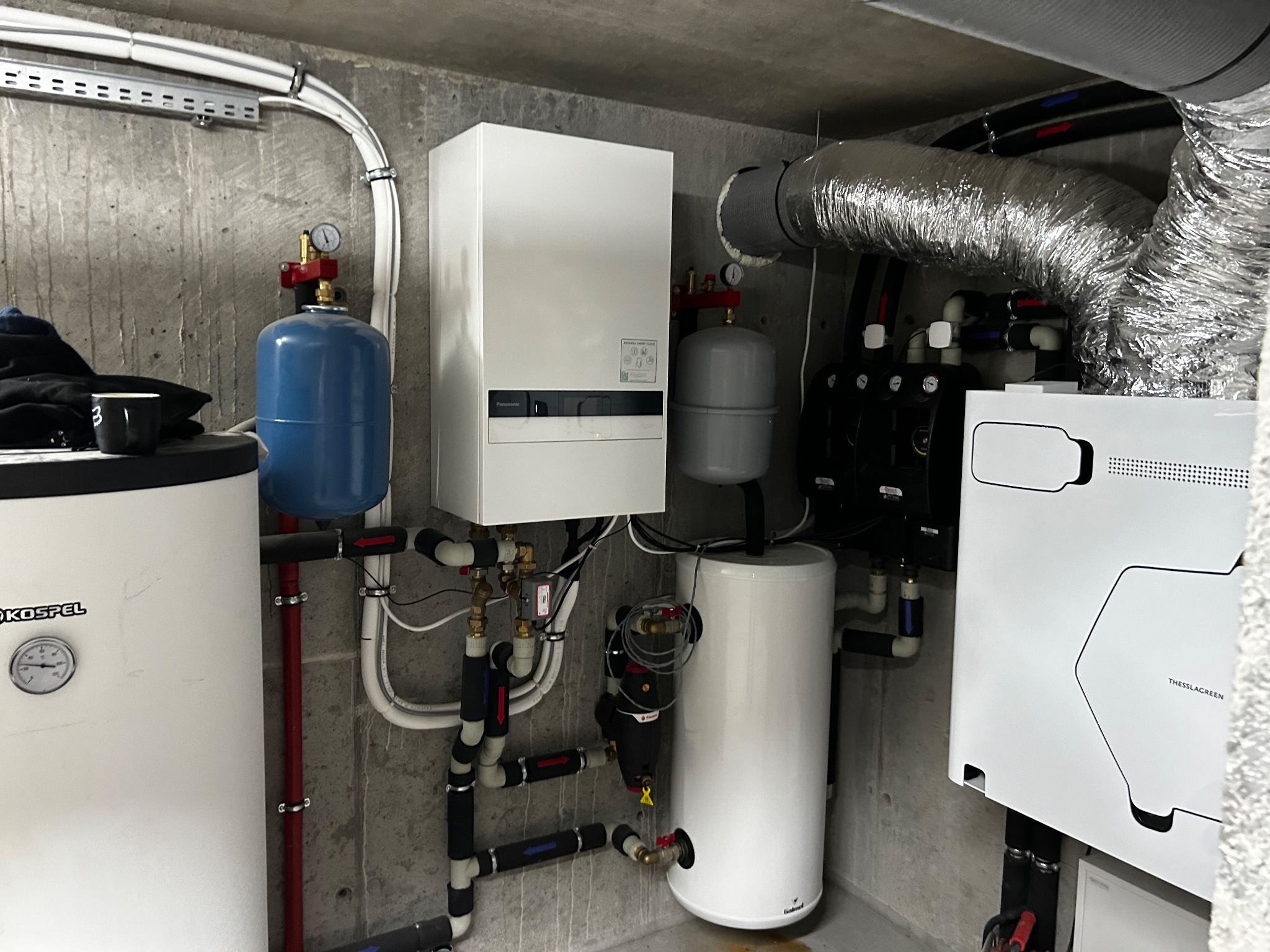 Kotłownia z piecem gazowym, zbiornikiem buforowym Kospel, instalacją hydrauliczną z rurami w izolacji i rekuperatorem Thessla Green, na tle surowego betonowego pomieszczenia.
