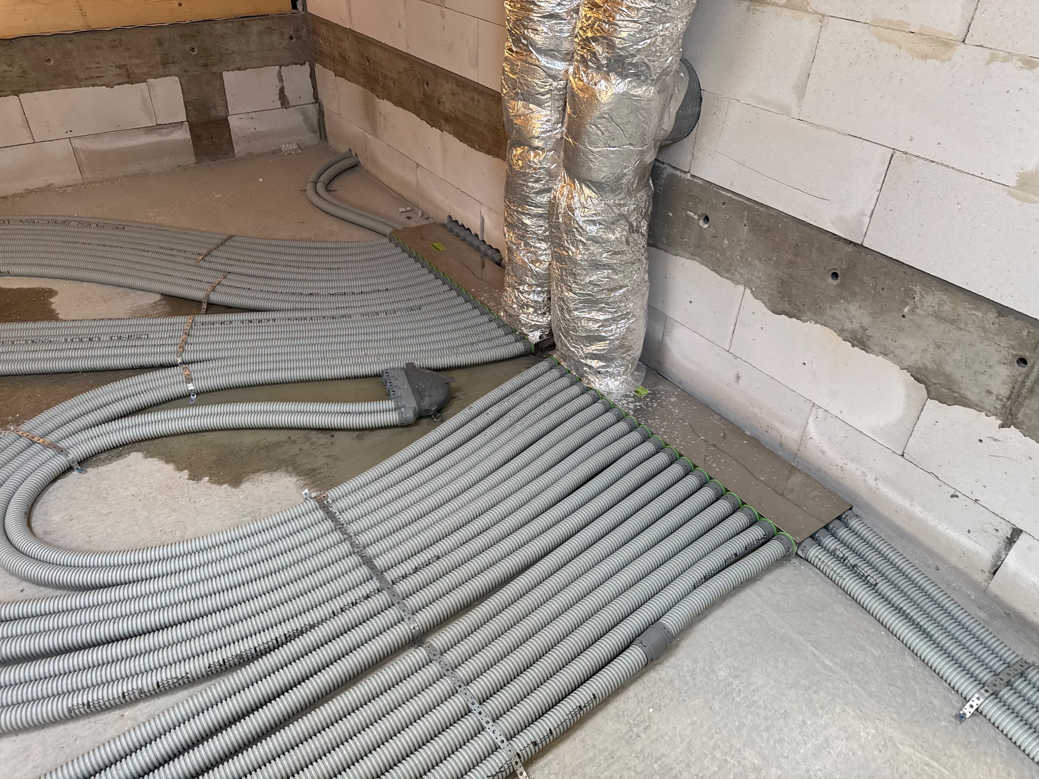 Szare, karbowane rury instalacyjne ułożone równolegle na podłożu w surowym wnętrzu. Widoczne mocowania i fragment ściany z bloczków.