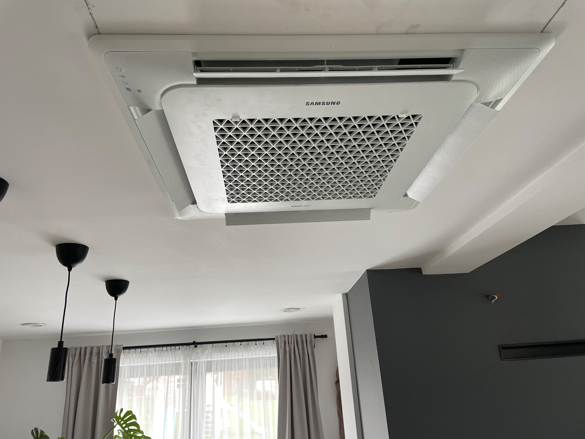 Sufitowy klimatyzator Samsung WindFree z widoczną kratką wentylacyjną w nowoczesnym wnętrzu, z czarnymi lampami wiszącymi w tle.