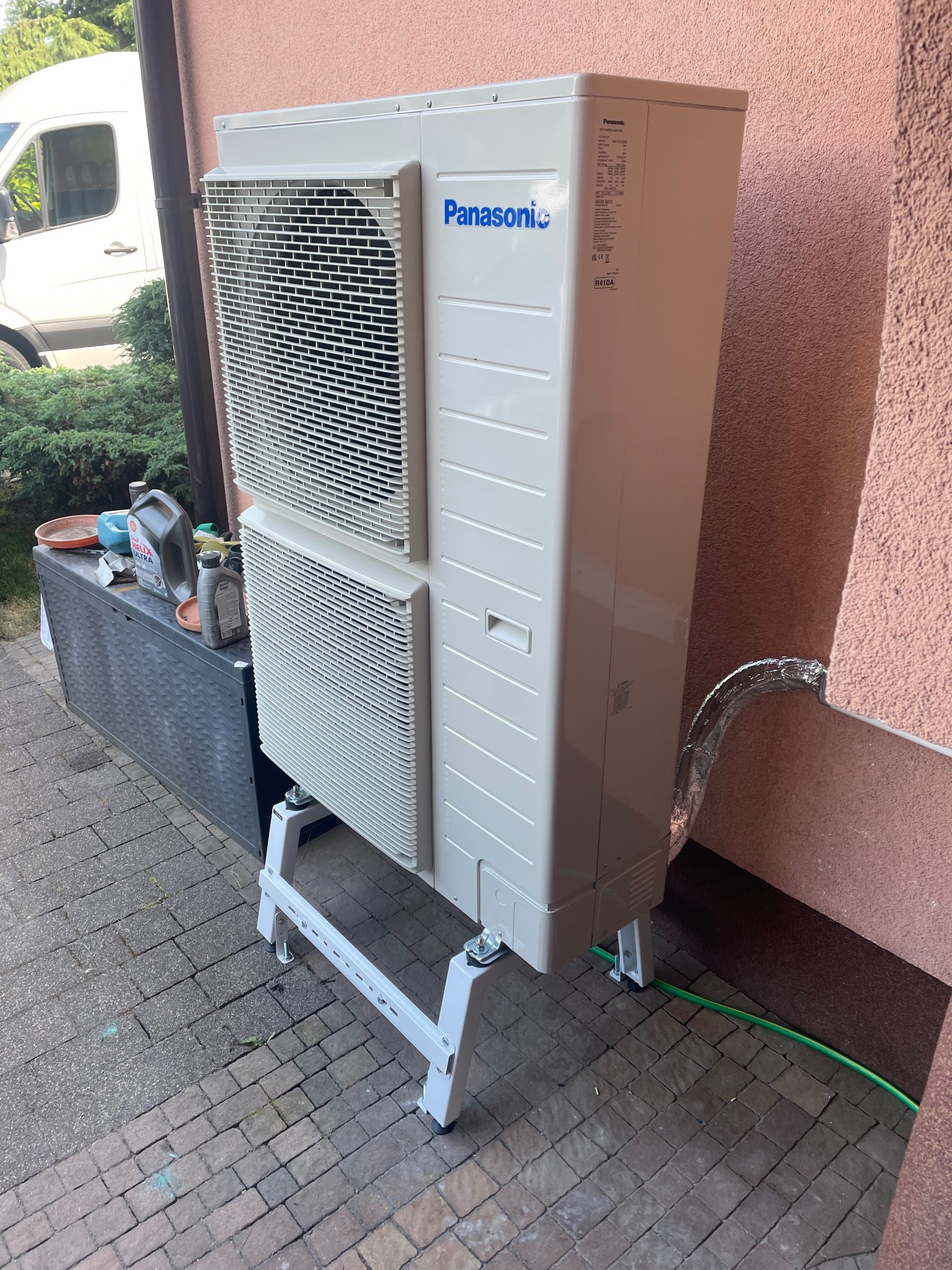Zewnętrzna jednostka klimatyzacji Panasonic zamontowana na białym stelażu przy ścianie budynku z widoczną izolacją rury. Obok skrzynia z narzędziami i olejami.