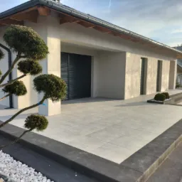 Nowoczesny taras z jasnych płyt, obrzeża z ciemnej kostki, ozdobne drzewko bonsai i białe kamienie w ogrodzie przed domem z szarymi roletami.