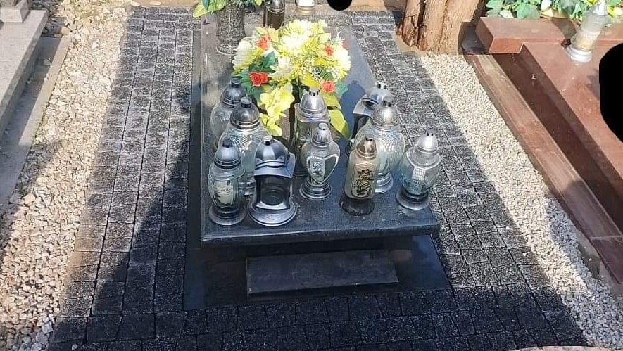 Ciemna kostka brukowa ułożona wokół nagrobka z licznymi zniczami i kwiatami, widok z góry.