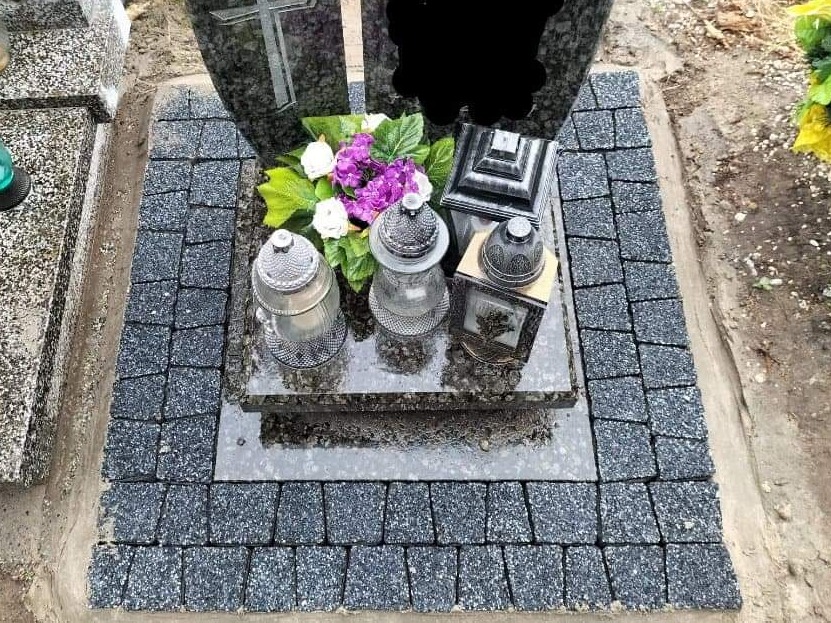 Obszar wokół nagrobka wyłożony drobną, szarą kostką, otaczający płytę z lampionami i kwiatami.