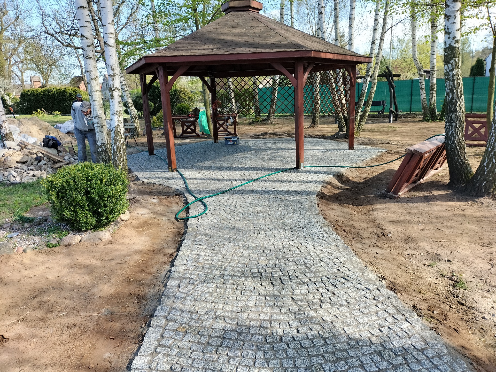 Ścieżka z kostki granitowej prowadząca do drewnianej altany ogrodowej, widoczny fragment ogrodu z drzewami i pracownik w tle.