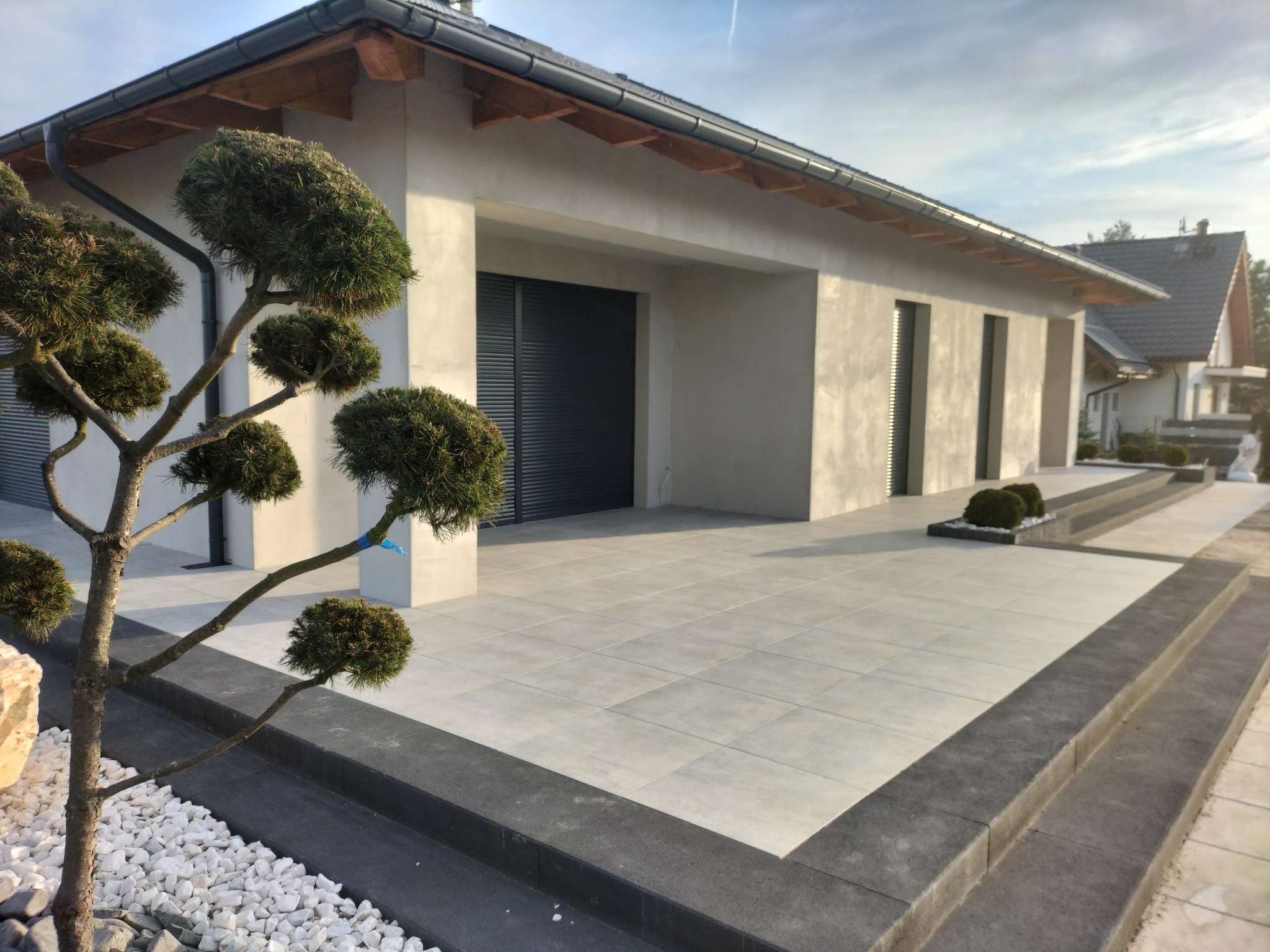 Nowoczesny taras z jasnych płyt, obrzeża z ciemnej kostki, ozdobne drzewko bonsai i białe kamienie w ogrodzie przed domem z szarymi roletami.
