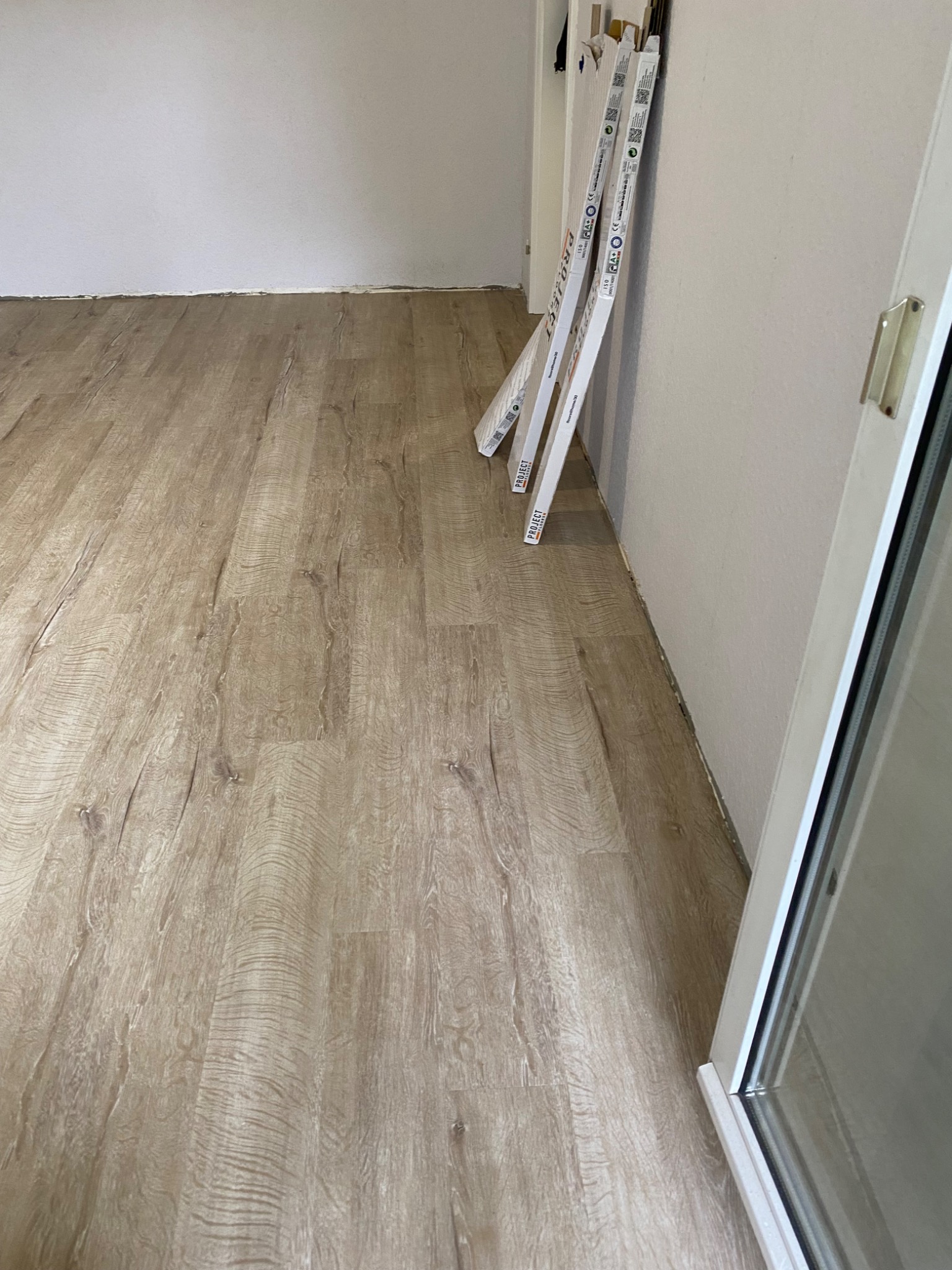 Ułożona podłoga z paneli w odcieniach drewna, przy ścianie oparte listwy przypodłogowe. Widoczny fragment białej futryny drzwi. Ujęcie z niskiego kąta.