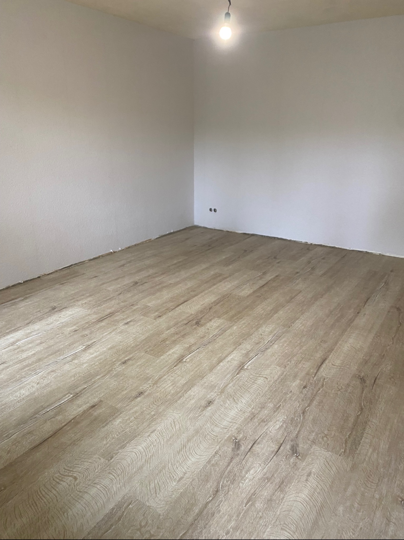 Ułożona podłoga z paneli w jasnym odcieniu drewna w pokoju w Starym Zawidowie. Widoczne ściany pomalowane na biało i surowy sufit z żarówką.