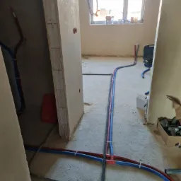 Wnętrze w trakcie remontu z widocznymi rurami instalacji wodnej i grzewczej, częściowo przymocowanymi do podłoża za pomocą opasek zaciskowych, z kartonem złączek w tle i oknem w oddali.